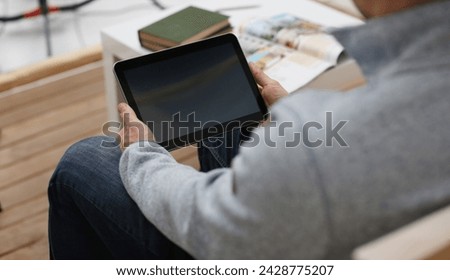 Male hand holds tablet pad in home setting while sitting on the couch engaged an internet surfing using application to press a finger on the display leisure listering music concept closeup.