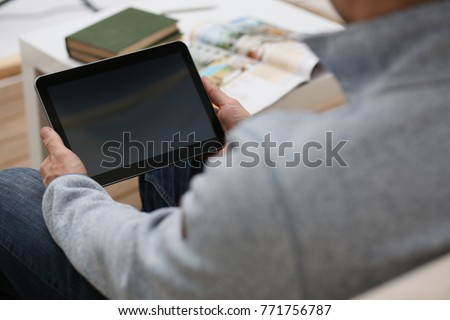 Male hand holds tablet pad in home setting while sitting on the couch engaged an internet surfing using application to press a finger on the display leisure listering music concept closeup.