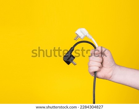 A male hand holds two electric plugs. No face, yellow background, copy space.

