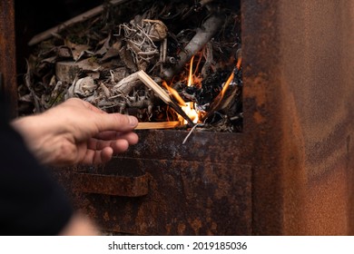 Male Hand Making A Fire With Small Wooden Branches In A Outdoor Rusty Metal Stove While Relaxing And Enjoying The Simple Life