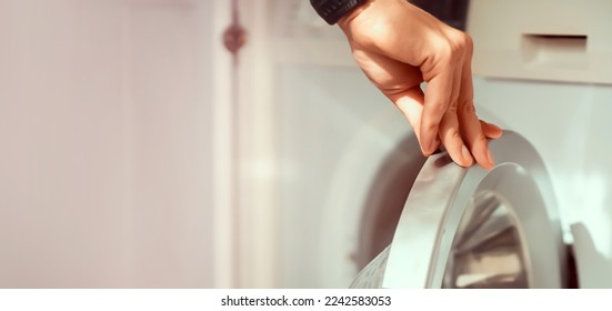 A male hand opens the door of a new washing machine in his modern, bright bathroom, close-up. The man washes clothes in the laundry. - Shutterstock ID 2242583053