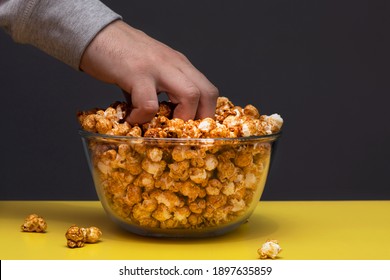 Male Hand Picks Up A Handful Of Crispy Popcorn In Caramel From A Glass Bowl On A Gray And Yellow Background