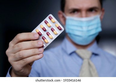 Male hand with pills, selective focus on fingers. Worried man in protective face mask and office clothes with medication in capsules at work
