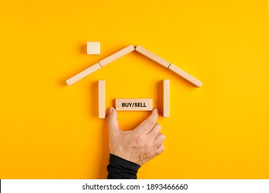 Male Hand Placing A Buy Or Sell Sign In A House Made Of Wooden Blocks On Yellow Background. Buying Or Selling A Hose Decision In The Real Estate Market. 