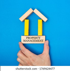 Male Hand Placing A Property Management In A House Made Of Wooden Blocks In A Conceptual Image Of Real Estate Market. Over Blue Background.