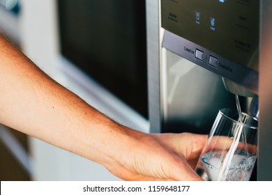 Male Hand Is Pouring Cold Water And Ice Cubes From Dispenser Of Home Fridge.