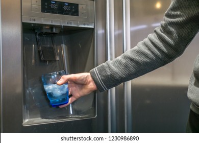 Male Hand Is Pouring Cold Water And Ice Cubes From Dispenser Of Home Fridge.