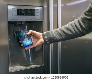 Male Hand Is Pouring Cold Water And Ice Cubes From Dispenser Of Home Fridge.