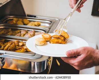 Male Hand Putting A Chicken Cutlet And Fried Potatoes  On A White Plate, Steam Table With Different Cooked Meats On A Background. Catering Or Breakfast Buffet At A Hotel.