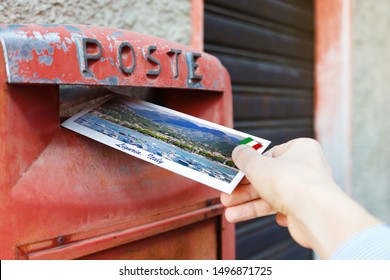 Male hand sends a postcard to a red mailbox in Liguria region, Italy. The postcard shows a Liguria coast, Sestri Levante city