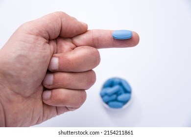Male hand showing a blue pill on a finger and in the background out of focus container full of blue pills