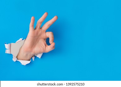 Male hand tearing through blue paper background creating ok gesture with hand and copy space for text.