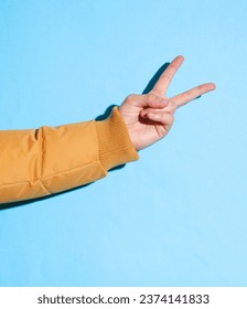Male hand in a warm winter jacket showing peace cymbol on a blue background