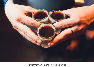 Male Hands Cheer With Glasses Of Shot Or Liqueur. Friends Drink Shot Or Liqueur And Cheers. Male Hands Cheer With Alcohol On Blurred Bar Background. Party And Toast Concept.