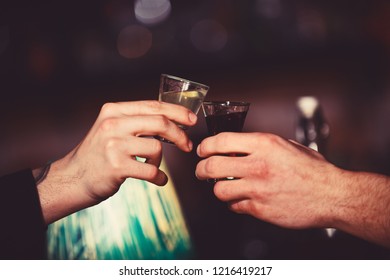 Male Hands Cheers With Alcohol On Blurred Bar Background. Friends Drink Shot Or Liqueur In Bar. Celebration And Alcoholic Beverages Concept. Male Hands Cheer With Glasses Of Shot Or Liquor