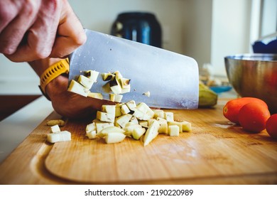 Male hands chopping peeled aubergine in cubes with a cleaver on wooden cutting board in a kitchen at daytime, preparing food for cooking, southern italian