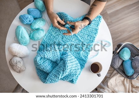 Male hands crochet handmade artwork use wool yarn and hook over table with coffee and potted plant top view closeup. Feminine man enjoy art hobby needlework handcrafted DIY thread sewing at home