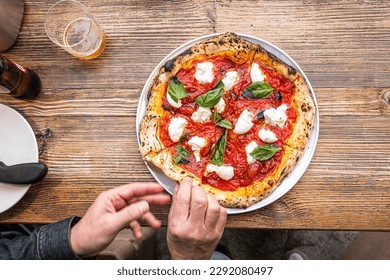 Male hands cutting a freshly baked Neapolitan Margherita Pizza on a rustic wooden table in a traditional Pizzeria. - Powered by Shutterstock