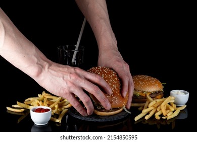 Male Hands Firmly Hold A Bitten Hamburger. Fresh Tasty Hamburger, French Fries, Cola On Black Glass With Reflection, Selective Focus, Top View. Fast Food, Junk Food For Quick Snack.