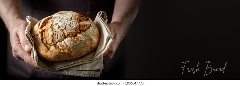 Male hands with fresh baked loaf rye wheat bread on a cotton towel. Dark background banner wirh copy space and text Fresh Bread