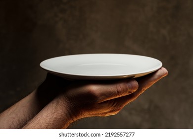 Male Hands Holding Empty Plate On Dark Background, Lack Of Food, Hunger And Crisis Concept
