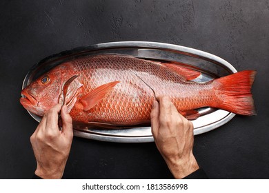 Male Hands Holding Fork And Knife Over Silver Tray With Giant Raw Fish On Black Stone Background. Overhead View, Flat Lay