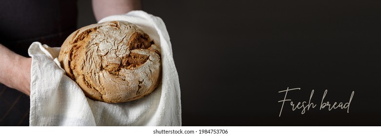 Male hands holding fresh baked loaf of rye wheat bread on a cotton towel. Dark background banner with copy space and text Fresh Bread - Powered by Shutterstock