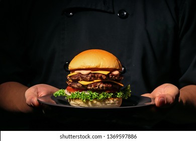 Male hands holding fresh delicious beef burger. Man in the black chef uniform serving homemade double patty burger with black angus beef, onion, tomato, cheese, lettuce, sauce on the black plate. - Powered by Shutterstock