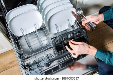 Male Hands Loading, Emptying A Dishwasher, Taking Out A Clean Fork From A Cutlery Basket. Household Chores With A Modern Kitchen Appliance. An Open Dishwasher With White Plates And Silver Cutlery.