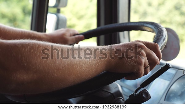 Male hands of lorry driver holds a big steering wheel
while driving a truck at summer day. Trucker riding to destination
at country road. Concept of logistics and transportation. Close up
Slow mo.