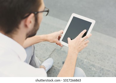 Male hands with tablet closeup, over shoulder shot. Businessman working on digital device with blank screen for copy space, sitting on stairs outdoors in urban area