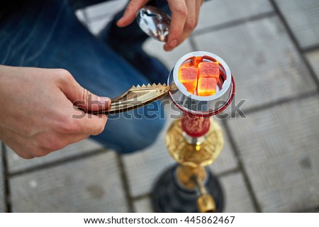 Male hands with tweezers and metal cap near hookah with live coals