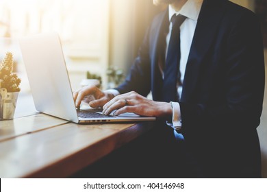 Male hands using laptop in modern coffee shop or loft, professional businessman in black suit working on new project with notebook computer while sitting at his office, flare light, blurred background