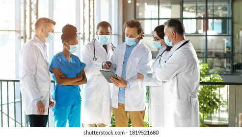 Male head of clinic standing with team of mixed-races doctors colleagues and using tablet device while watching on screen. Group of multi ethnic medics, men and women talking and having discussion.