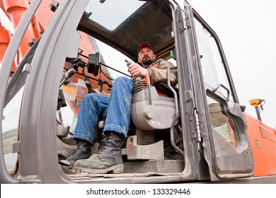 A Male Heavy Equipment Operator Sitting In The Cab Of A Track-hoe At A Construction Site On A Cloudy Winter Day.