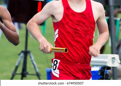 A male High School runner is racing in a relay race wearing a red uniform and carrying a gold baton.