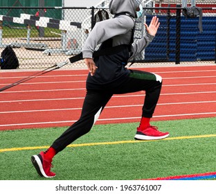 A Male High School Runner Wearing A Protective Face Mask Is Dragging A Sled With Weight Across A Green Turf Field During Strength And Agility Practice.