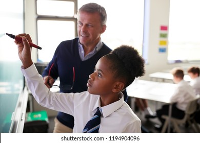 Male High School Teacher With Female Student Wearing Uniform Using Interactive Whiteboard During Lesson