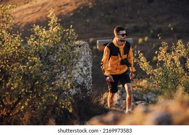 Male Hiker With Backpack And Digital Camera Walking On Hill During Sunset, Traveler Man In Solo Trip