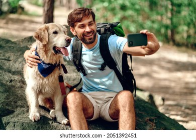 A male hiker with a backpack and a labrador dog makes a selfie on the phone while sitting on a stone in the forest. Camping, travel, hiking. - Powered by Shutterstock