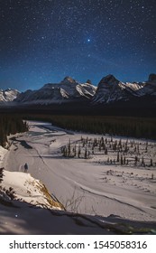 Male Hiker In Blue Jacket Standing At Goats And Glaciers At Night In Winter. Jasper National Park, Icefields Parkway, Canadian Rocky Mountains, Alberta, Canada. Wild, Dark Starry Sky, Portrait Layout