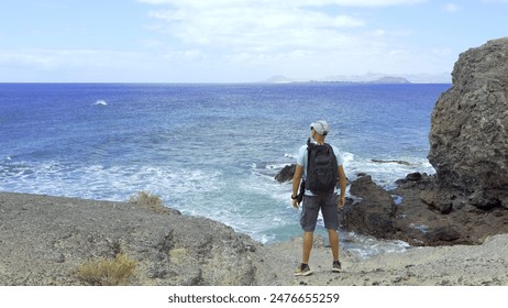 A male hiker contemplates the ocean from the lava rock shore. The man is a young adult and Hispanic. The boy is photographed standing and from behind. Freedom and summer travel concept - Powered by Shutterstock