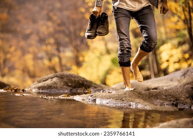 Male hiker crossing the stream barefooted walking by the creek with his shoes in hand adventure travel hiking trail forest.	