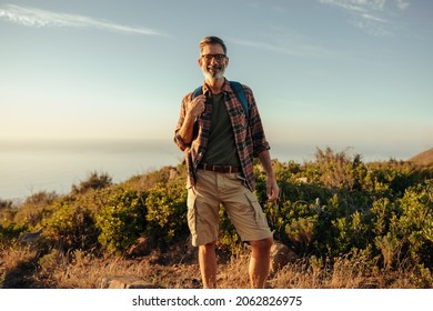 Male hiker looking at the camera on a hilltop. Happy mature hiker smiling while standing on top of a hill with a backpack. Adventurous male backpacker enjoying a hike near the sea. - Powered by Shutterstock