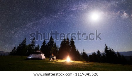Male hiker sitting alone near tourist tent at bonfire on grassy valley, enjoying night blue starry sky with full moon, Milky way, pine trees forest on background. Tourism and mountain camping concept