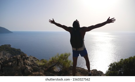 Male Hiker Standing On Edge Mountain With Raising Hands And Admiring Scenic Seascape. Man With Backpack Actively Resting During Trip Enjoying Freedom On Hill Top. Beautiful View At Background. Slow Mo