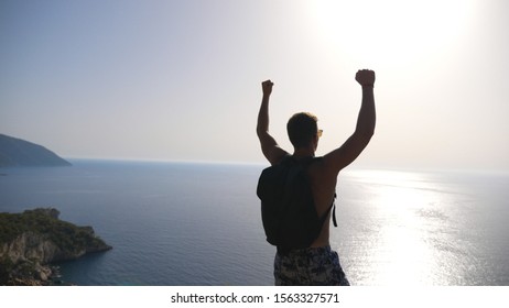 Male Hiker Standing On Edge Mountain With Raising Hands And Admiring Scenic Seascape. Man With Backpack Actively Resting During Trip Enjoying Freedom On Hill Top. Beautiful View At Background. Slow Mo