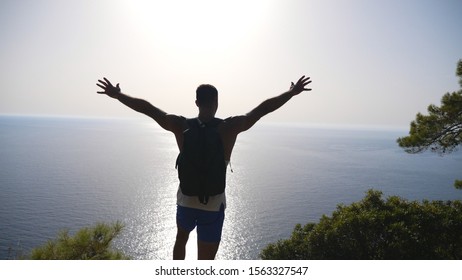 Male Hiker Standing On Peak And Victoriously Raising Hands Rejoicing Success. Young Man With Backpack Actively Resting During Trip Enjoying Freedom At Hill Top. Scenic Seascape At Background. Slow Mo