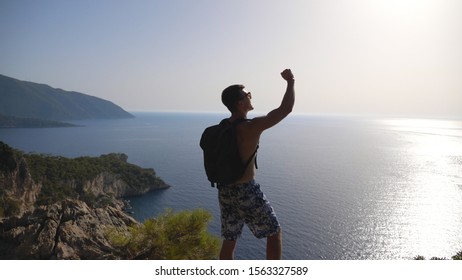 Male Hiker Standing On Peak And Victoriously Raising Arm Rejoicing Success. Young Man With Backpack Actively Resting During Trip Enjoying Freedom At Hill Top. Scenic Seascape At Background. Slow Mo