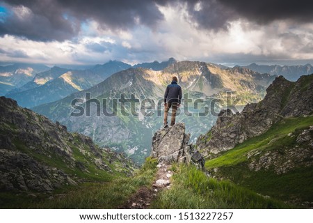 Male hiker standing on a rock above deep valley in front of scenic panorama of high mountains with dark clouds above him.